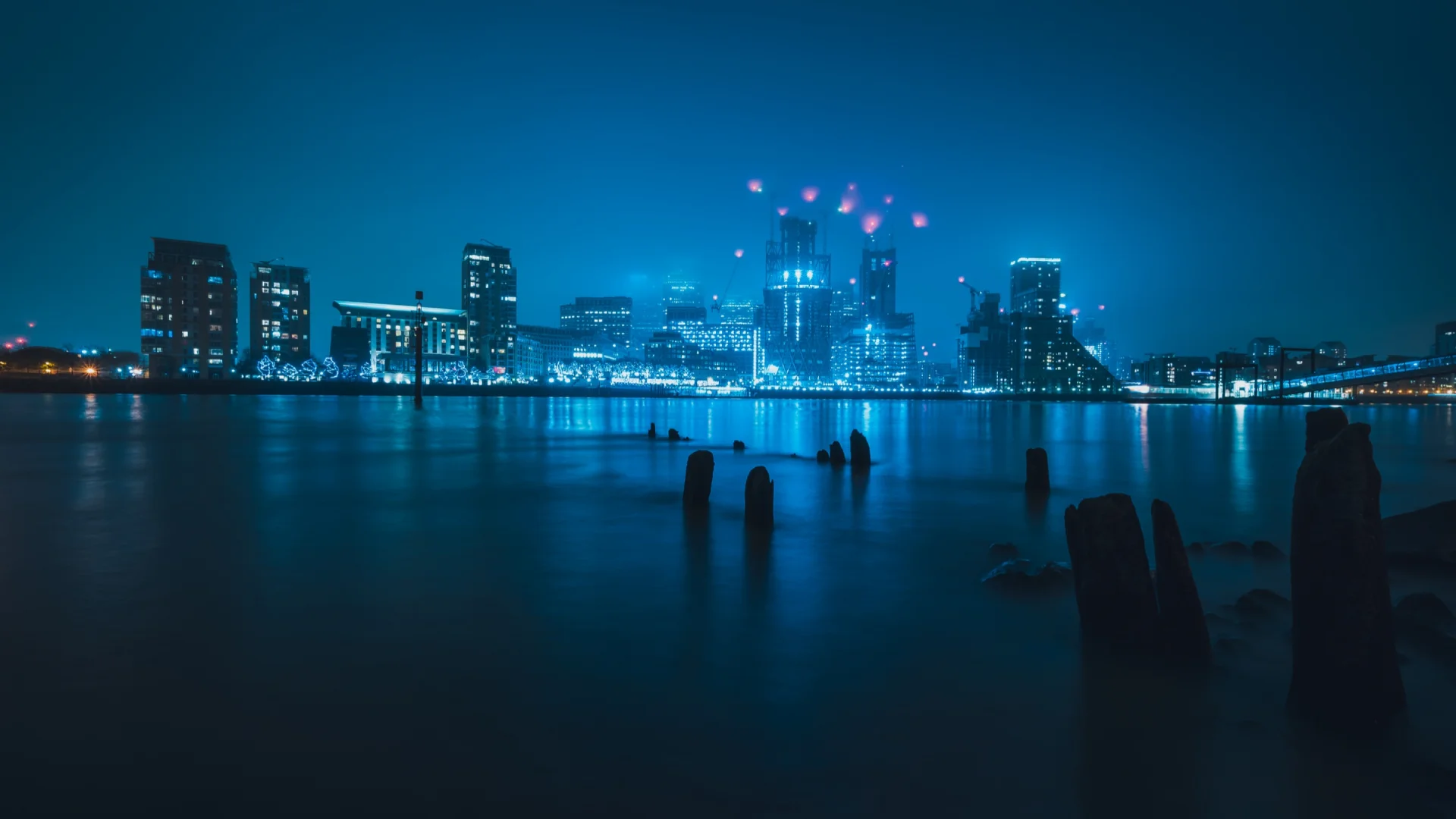 La silueta de Londres de noche sobre el agua, con fuegos artificiales encima de las torres y postes de madera en primer plano.