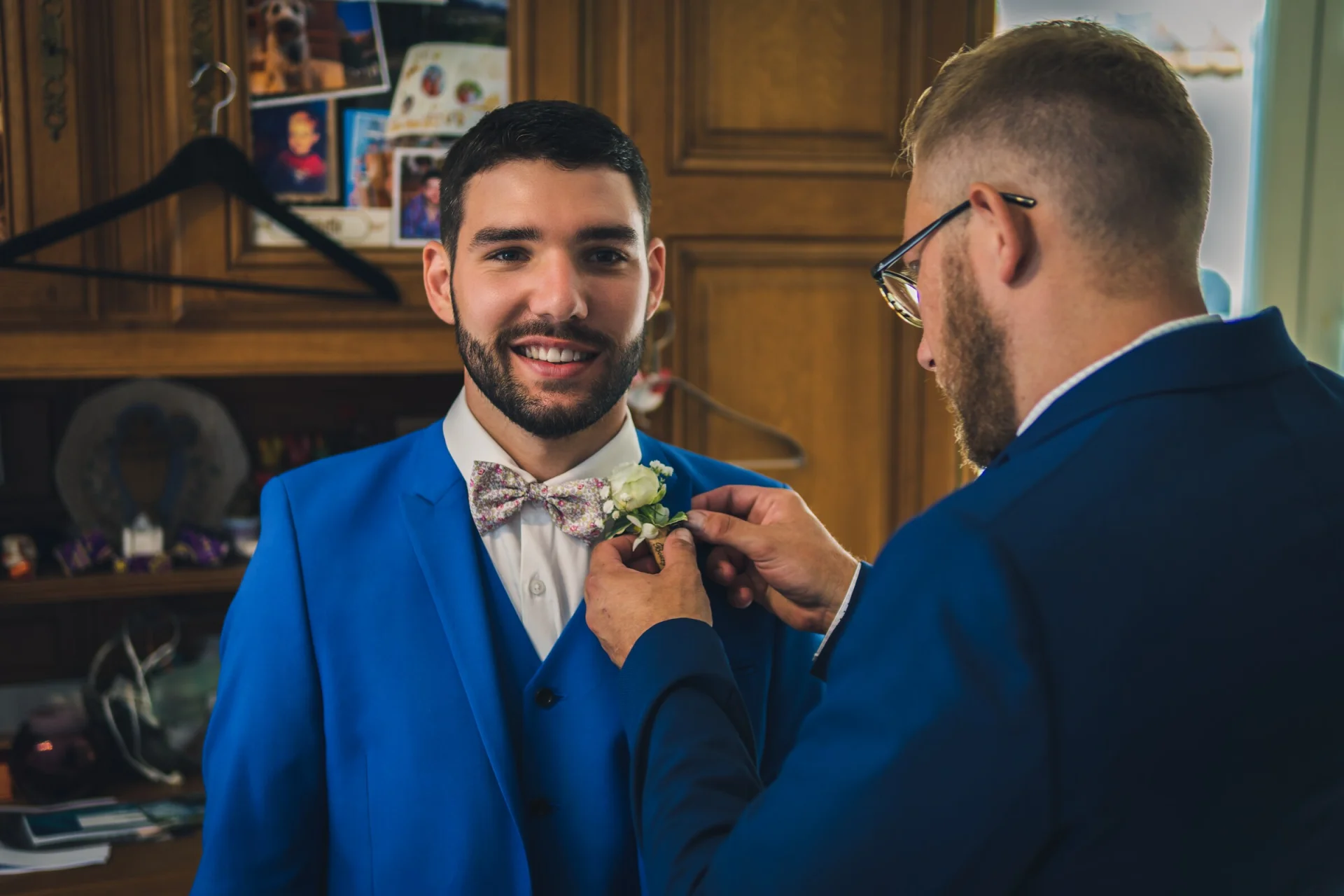 Un témoin ajuste la boutonnière du marié, en costume bleu.