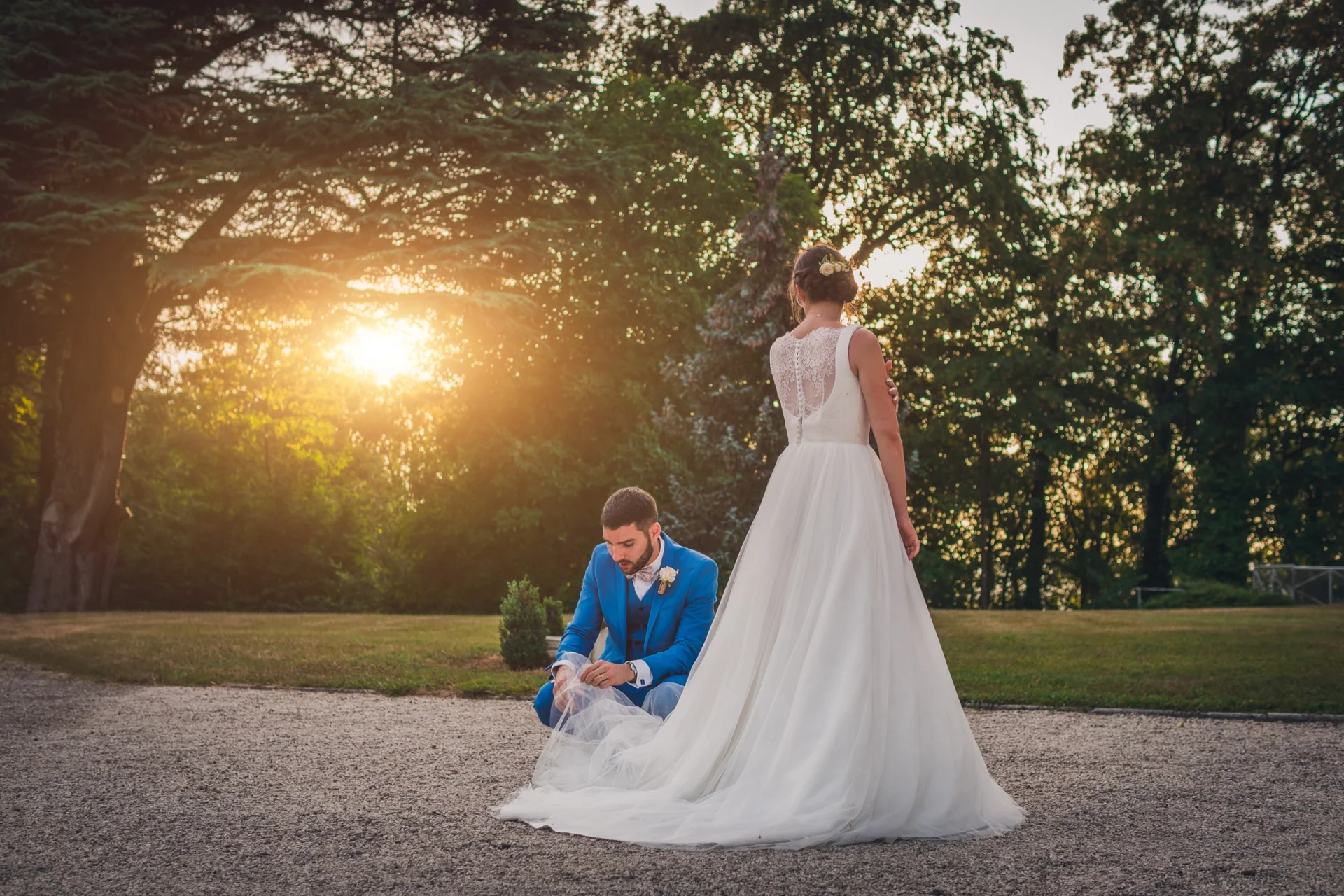 Una novia de espaldas y el novio arrodillado ante ella, bajo grandes árboles al atardecer.