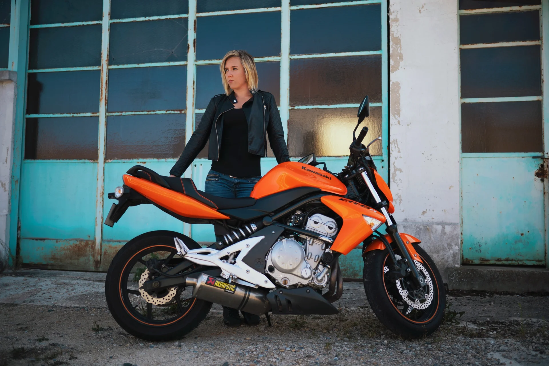 Una mujer sentada en una moto naranja ante la fachada de un taller con cristales turquesa.