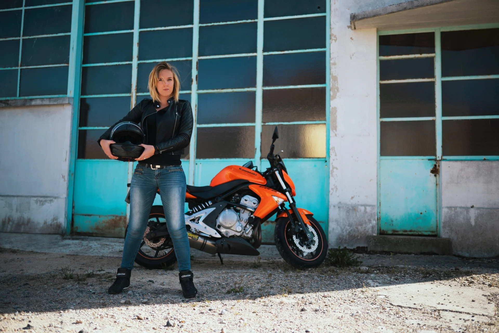 Una mujer de pie ante una moto naranja, con el casco en la mano, en una mancha de sol.