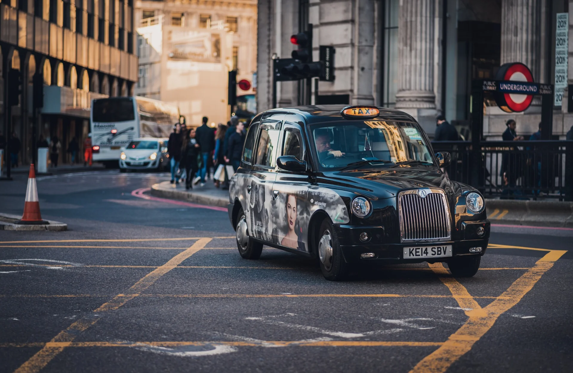 Un taxi noir londonien traverse un carrefour, l'entrée du métro derrière lui.