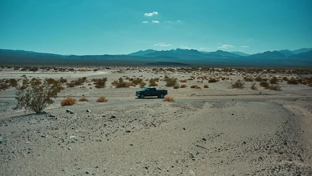 Un pick-up minuscule sur une piste du désert, les montagnes bleues à l'horizon.