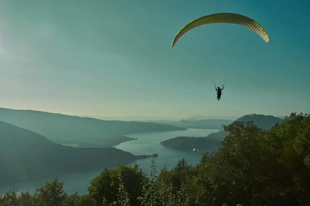 Un parapente amarillo pasa cerca del despegue, con el lago de Annecy y las montañas detrás.